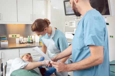 Smiling woman lies on a table, shaking hands with male doctor as female nurse takes her blood pressure.