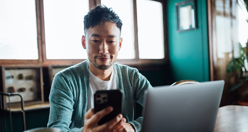 A man looking at smartphone while working on laptop computer in home office.
