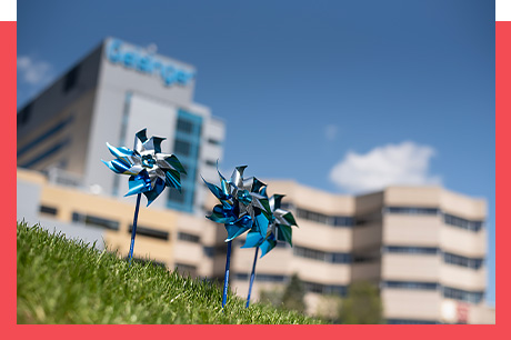 Pinwheels in focus with blurred hospital building in the background under a clear sky.