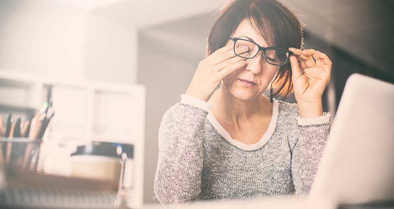 an image of a woman rubbing her eyes while sitting in front of a laptop